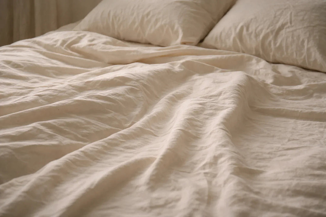 Lit minimaliste aux draps sable doux légèrement froissés avec deux oreillers posés, ambiance calme et naturelle évoquant le confort nocturne et le sommeil réparateur.