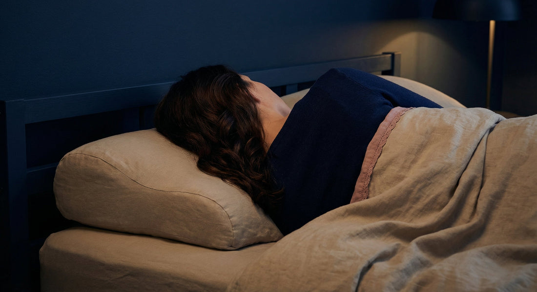 Femme aux cheveux châtains ondulés allongée sur le côté dans un environnement nocturne calme, reposant sur un oreiller ergonomique en lin beige, linge doux et lumière basse.