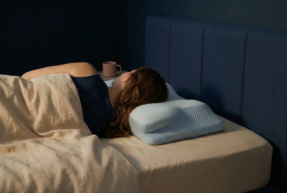 Femme aux cheveux châtains allongée sur le côté sur un lit double, tête posée sur un oreiller ergonomique, dans une chambre nocturne aux tons bleu nuit et sable doux.